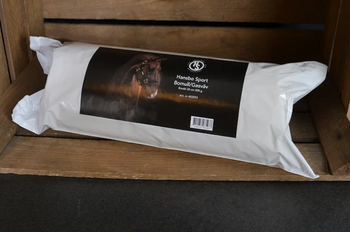 Ett vitt paket märkt Bomull/Gasväv med en hästbild på en trähylla.