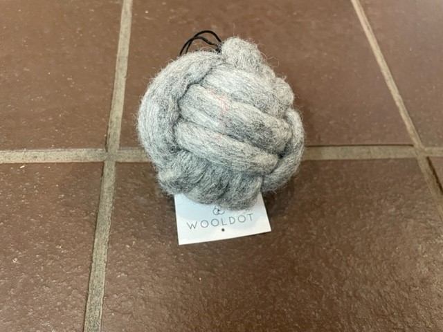 En 8 cm stor boll med hundknut ligger på ett brunt klinkergolv med en WOOLDOT-tagg fäst.