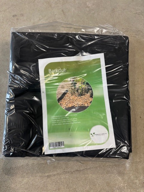 En förpackad svart trädgårdstäckning märkt Barkduk 1,6x10m 16kvm med mulch och växter bild.