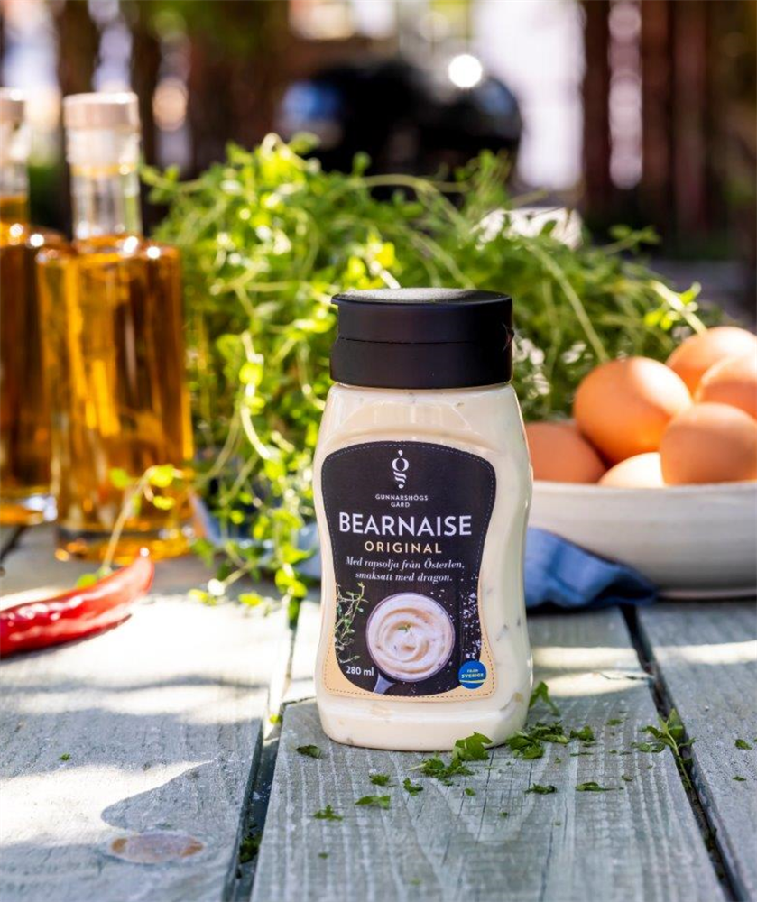 Bearnaisesås original 280 ml på ett bord med ägg, örter och oljeflaskor i bakgrunden.