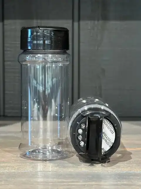 Två genomskinliga 100 ml KRYDDBURK-burkar med svarta lock - en stående och en liggande på sidan.