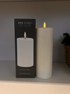 LED WAX CANDLE D7,5 H20 i vitt visas bredvid sin låda på en hylla.
