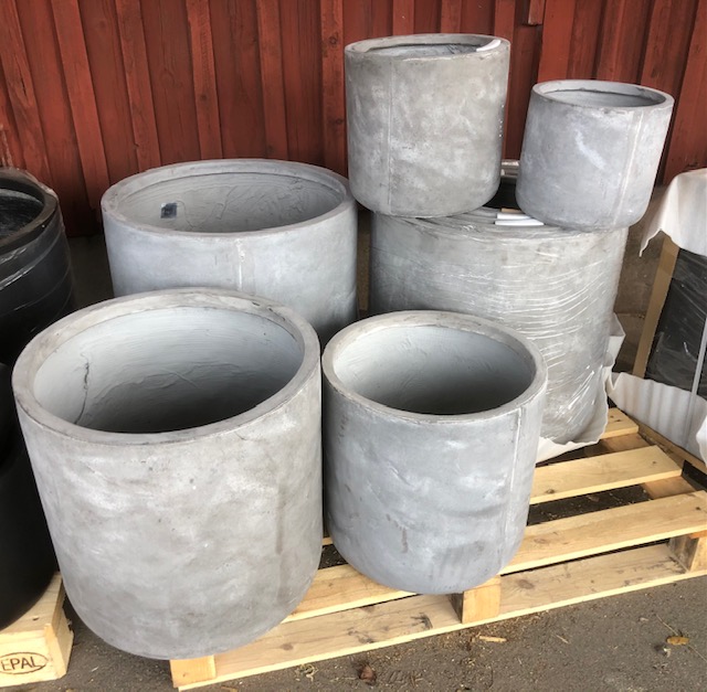 Grå betongcylindrar i olika storlekar staplade på en träpall vid en röd trävägg.