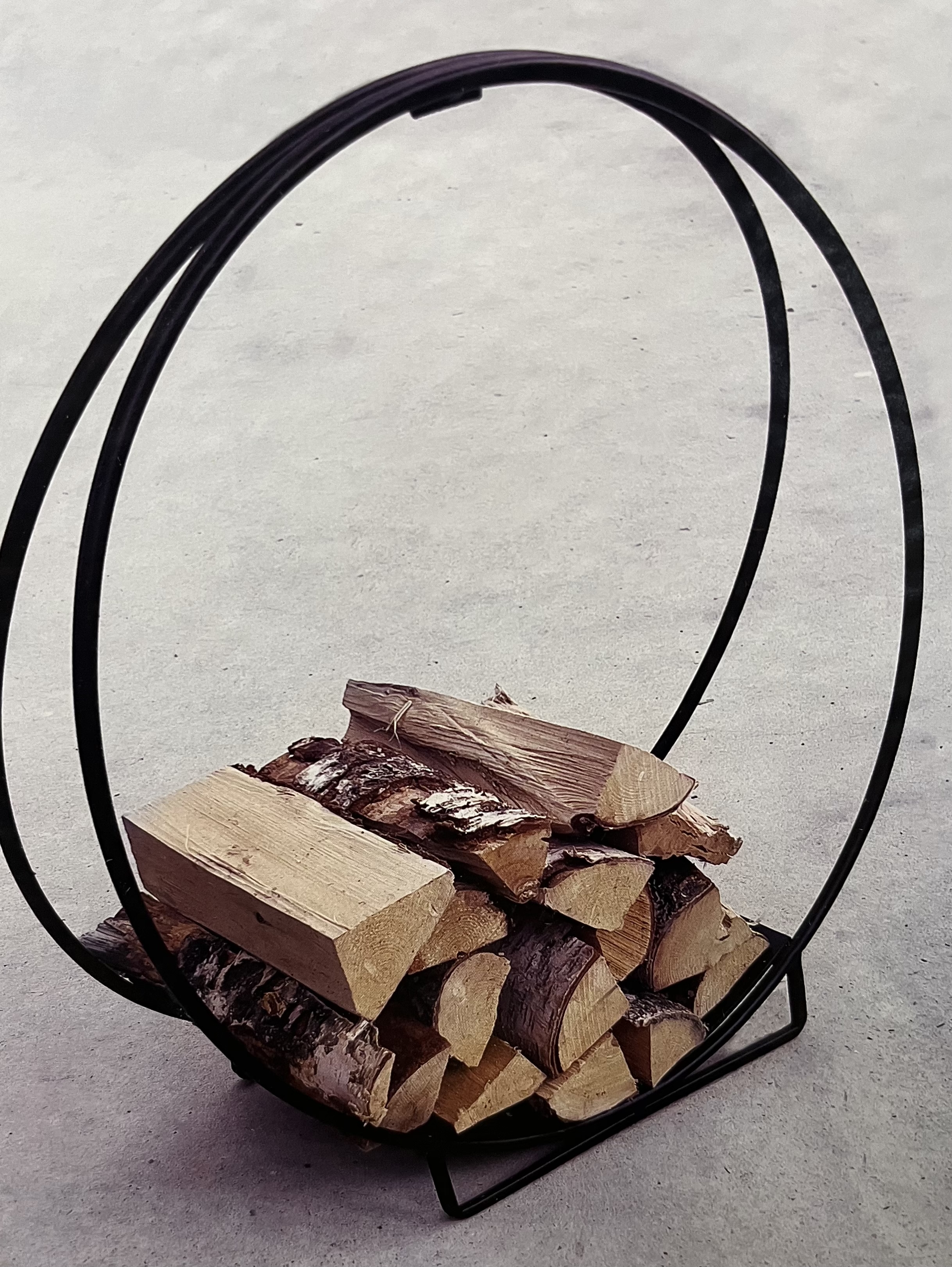 Vidställ 71X70X28,5 rymmer staplad ved i en rund vedhållare i svart metall på grå botten.