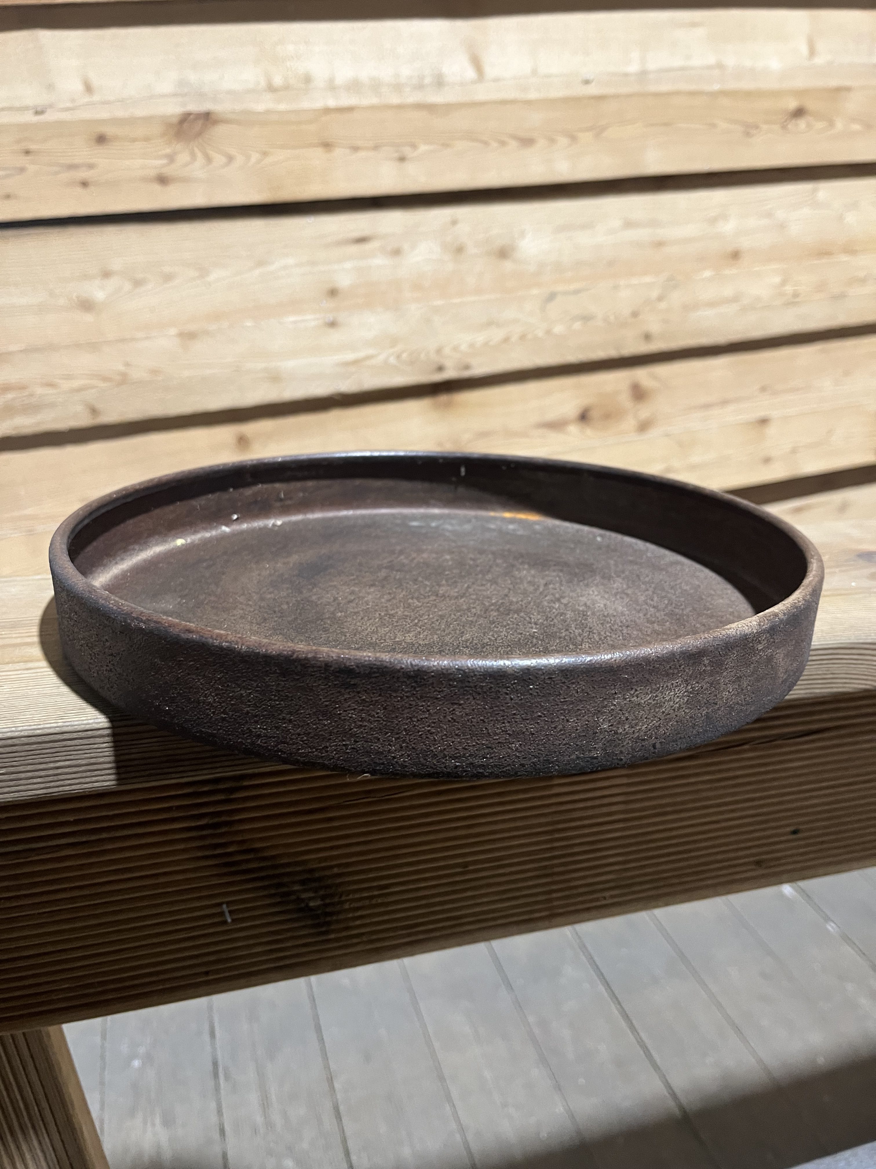 Den åldrade rostbrickan Fat cylinder 28,5x28,5x3,5 vilar på en träbänk i solljus.