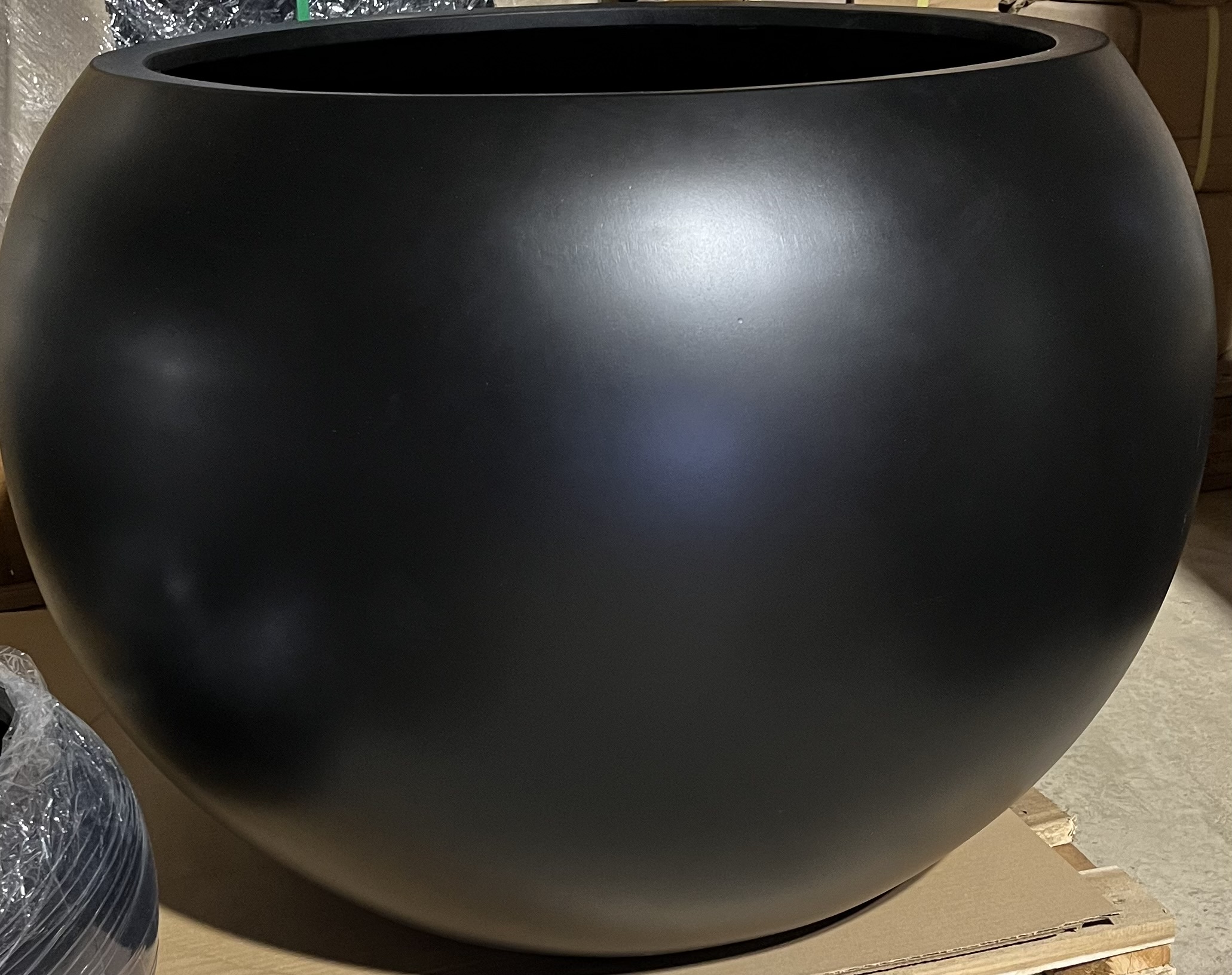 Kruka poly black 82x57, en stor rund matt svart kruka, står på en kartongyta inomhus.