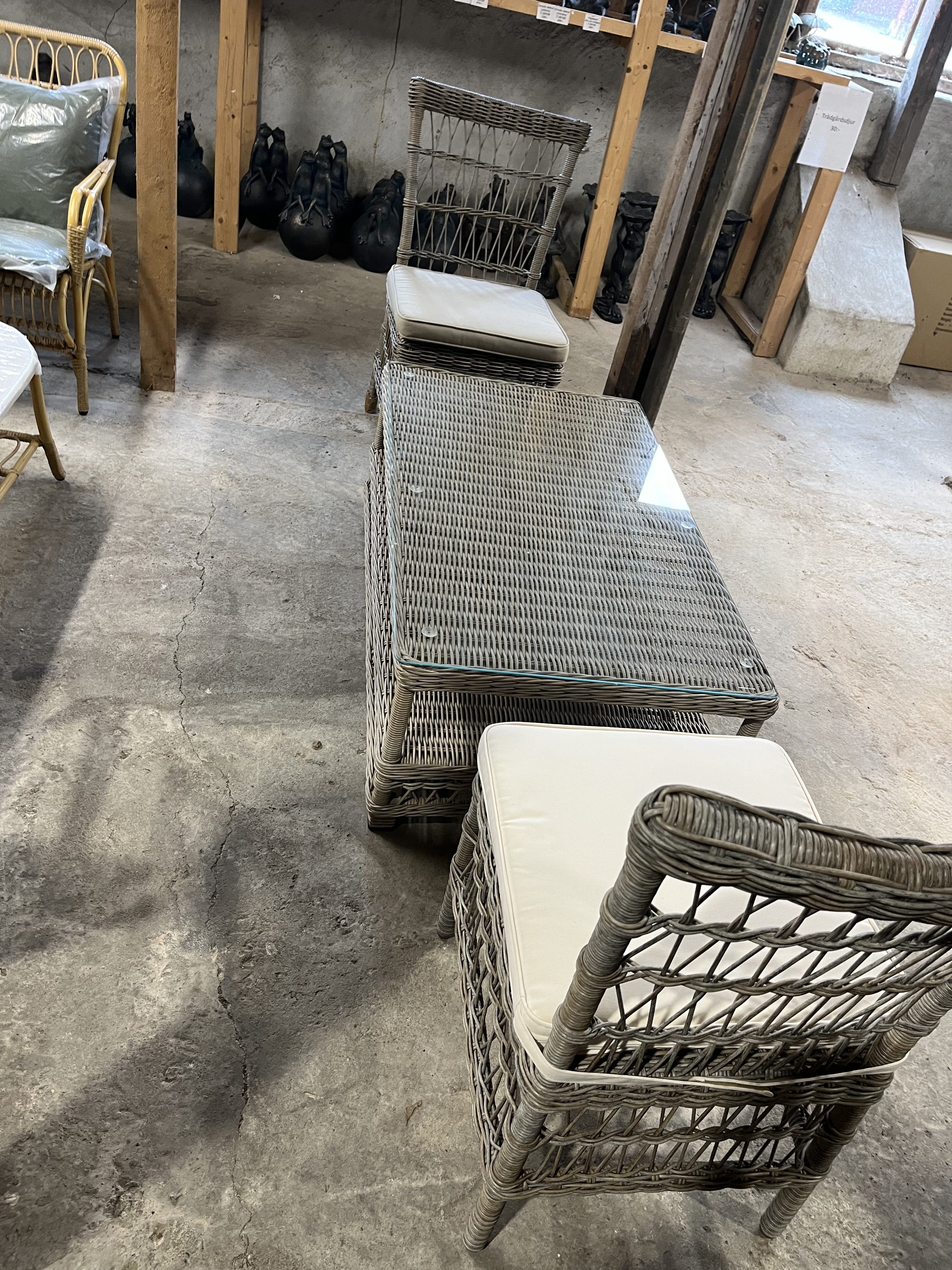 Två Stol 1 korgstolar och ett bord med glasskiva och vita dynor på ett betonggolv.