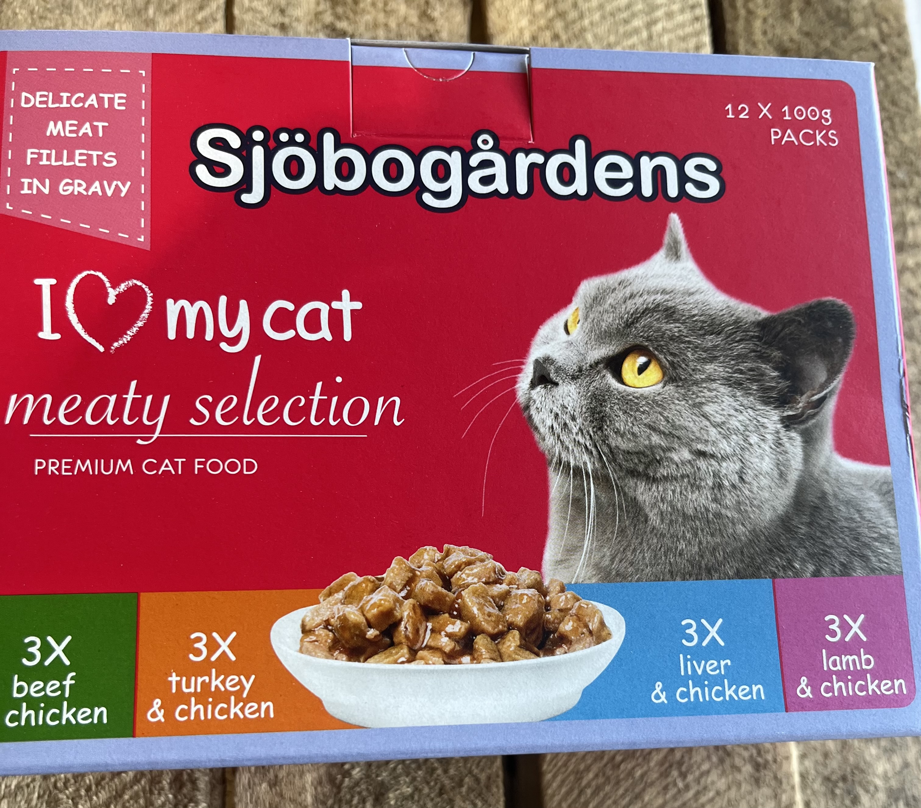 Kattmat 12p Köttmeny Fancy meat 1,2kg box med grå katt som tittar på en skål med kött.