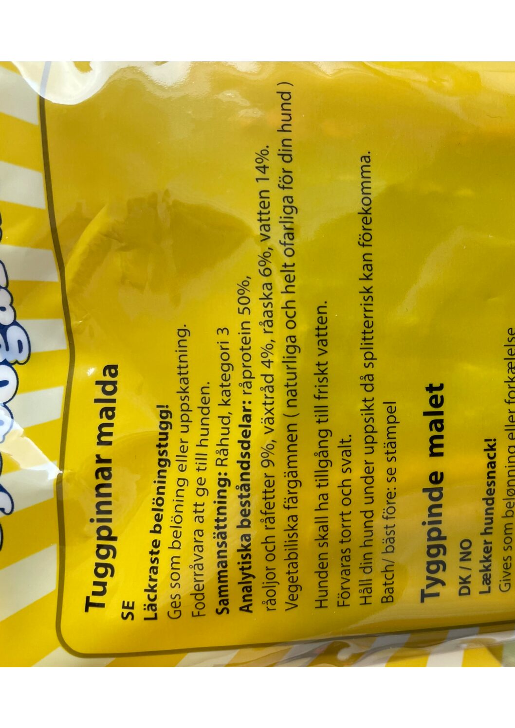 Gul förpackning med svensk och dansk text som beskriver tuggpinnar för hundar, mot en randig bakgrund.