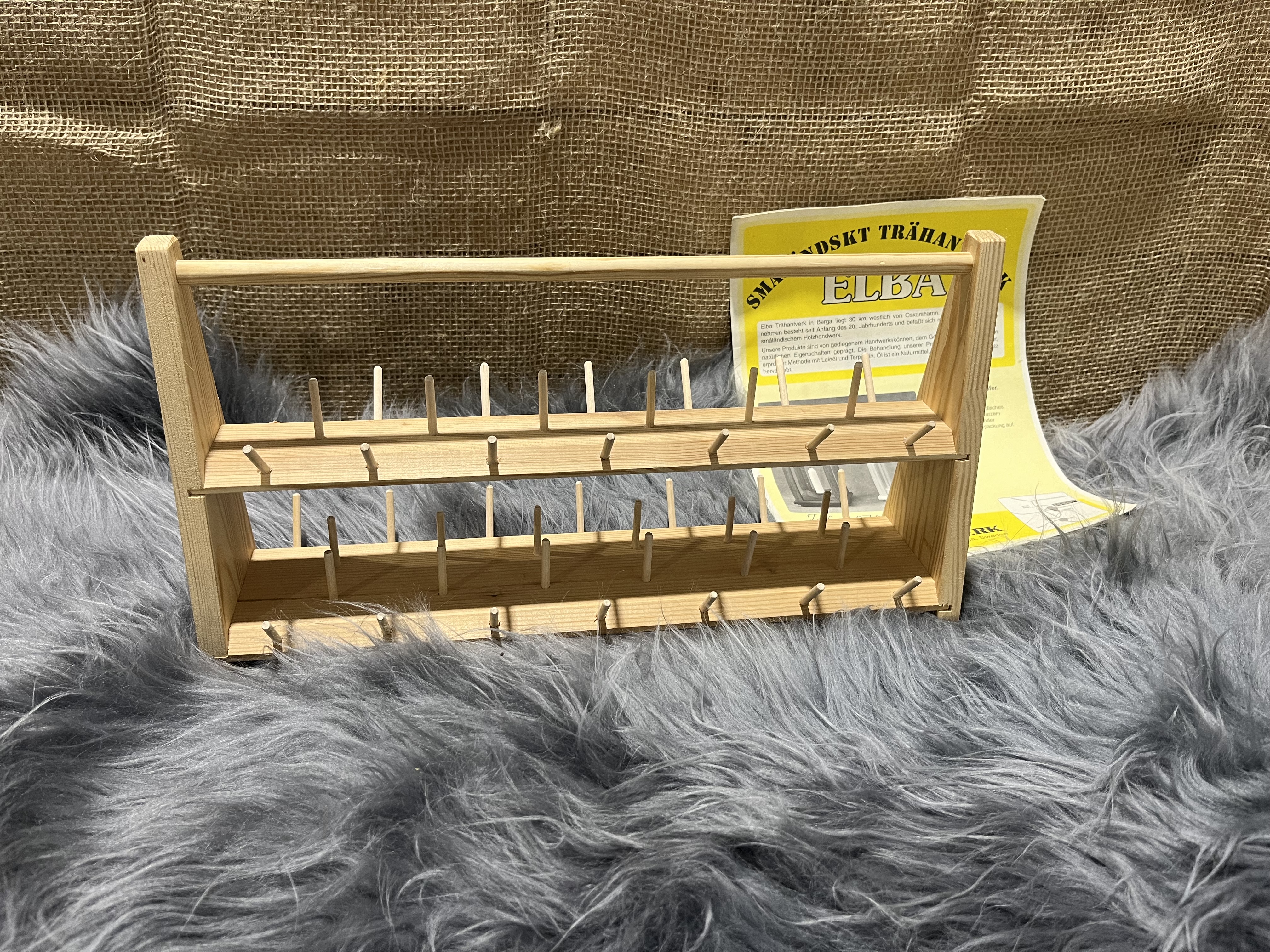 Trä trådrullsställ med två pinnrader på grå fuskpäls, gul broschyr i bakgrunden.