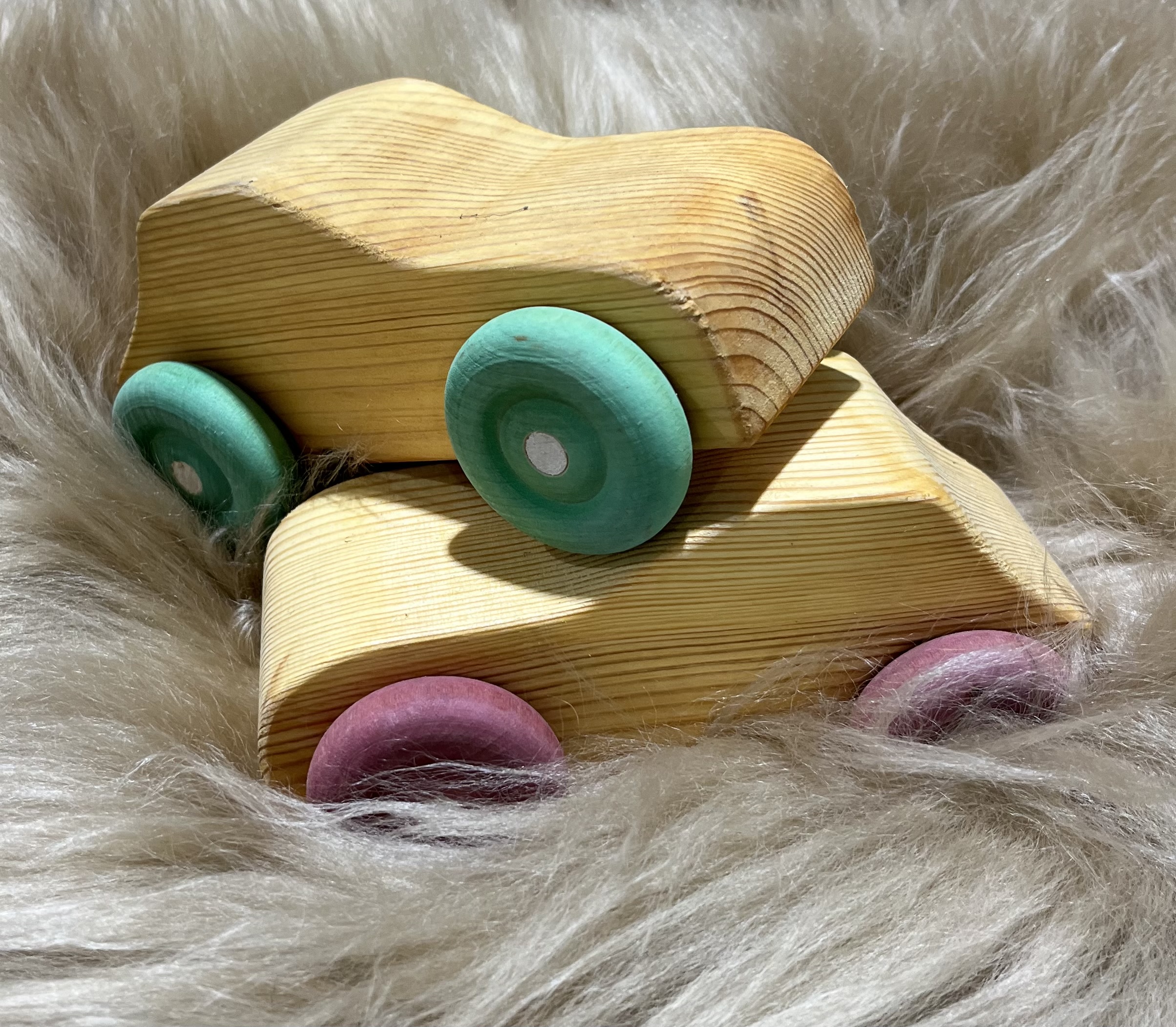 Två Trä bil leksaksbilar i trä med pastellfärgade hjul på ett mjukt, fluffigt underlag.