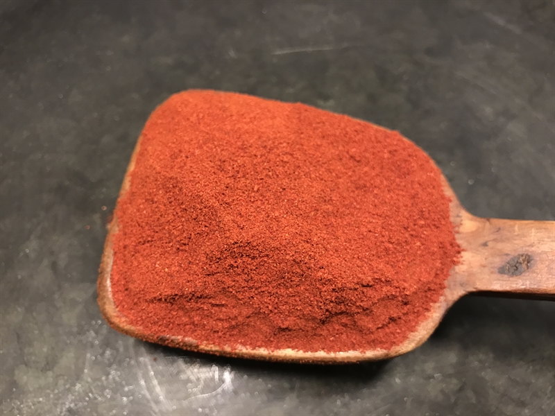 En träsked fylld med Paprika Mild-pulver på en mörk yta.