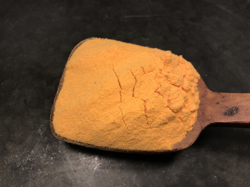 En träsked håller i Popcorn Krydda orange pulver mot en mörk bakgrund.