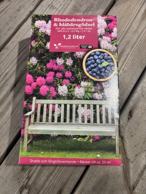 KRAV Rhodo&Blåbärsgödsel 8-2-5 1,2L på trä med blommor och en bänk i bakgrunden.
