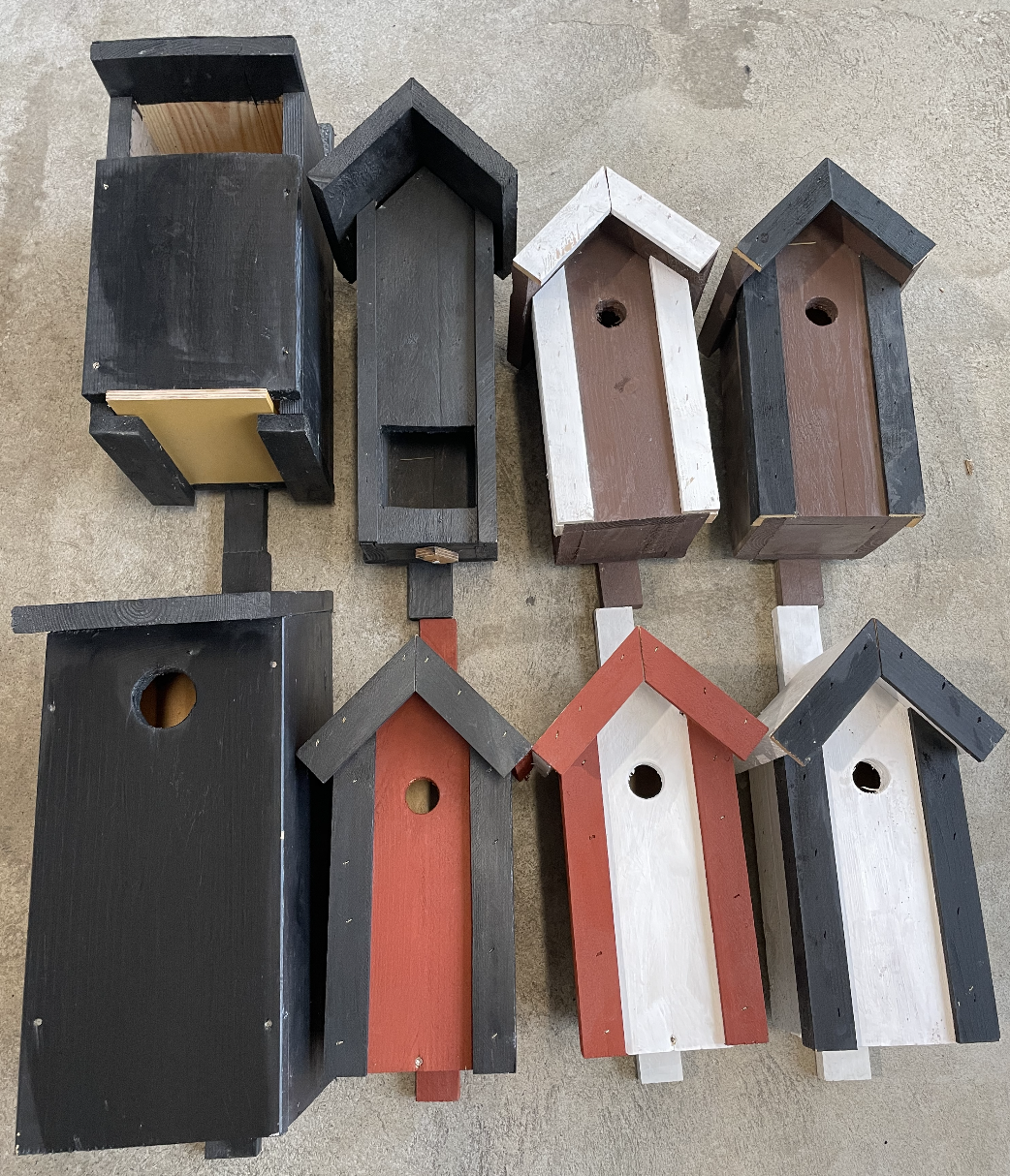 Åtta fågelholkar i olika former och färger placerade på en betongyta.