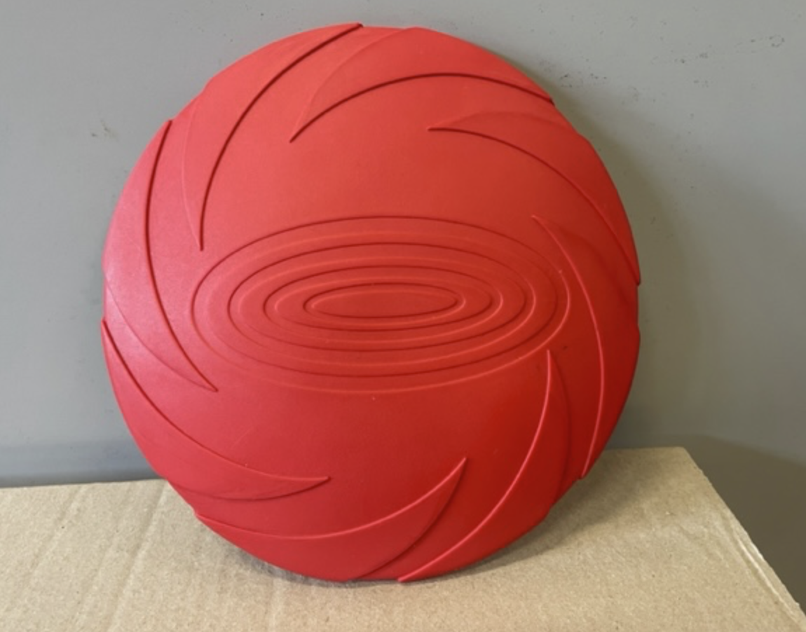 Mjuk FRISBEE: Röd, cirkelformad med spiralmönster, placerad på kartong vid en grå vägg.