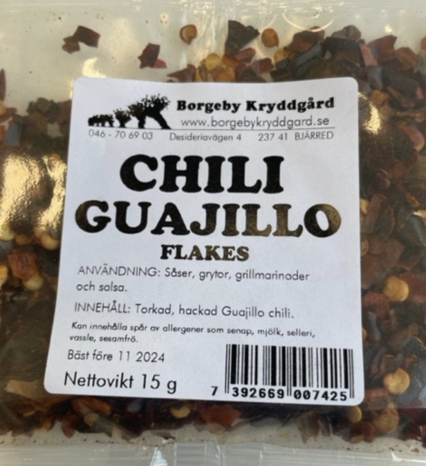 En genomskinlig påse med CHILI GUAJILLO FLAKES (15 g) med en vit produktetikett.