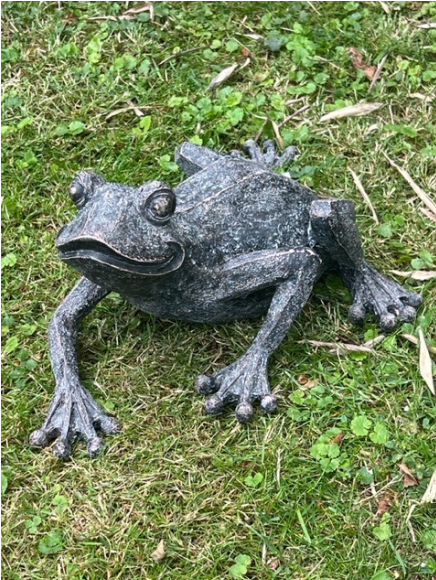 En grodstaty i sten sitter i gräset omgiven av små gröna blad och växter.