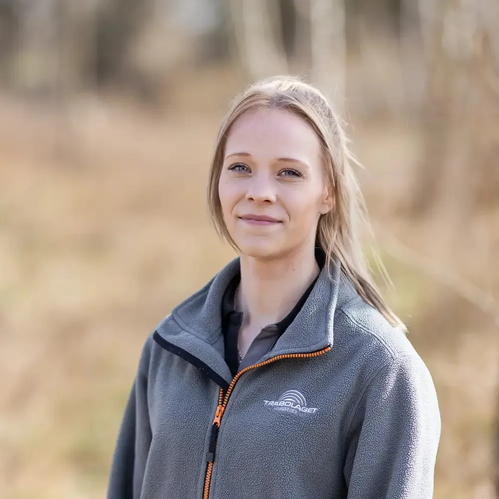 En kvinna i grå jacka står utomhus och ler mjukt mot en oskarp naturlig bakgrund.