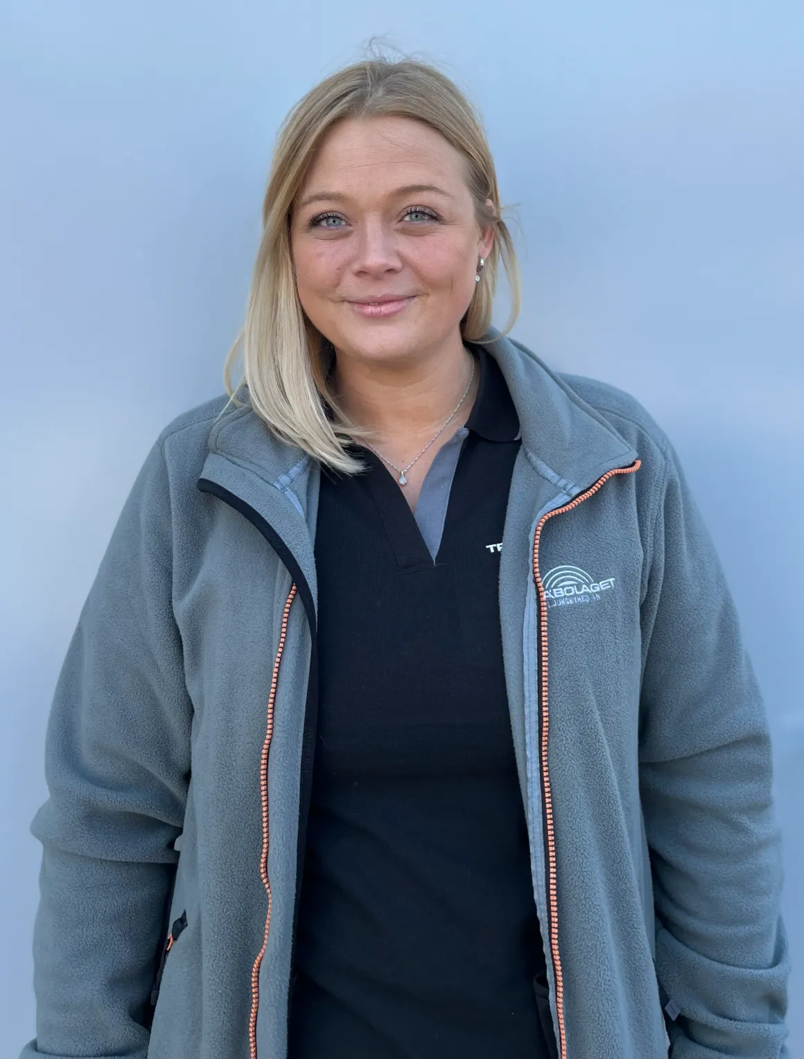 En kvinna i grå jacka och svart skjorta står mot en ljusblå bakgrund och ler.