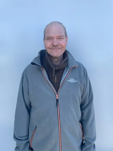 En man i grå kavaj ler när han står framför en ljusblå bakgrund.