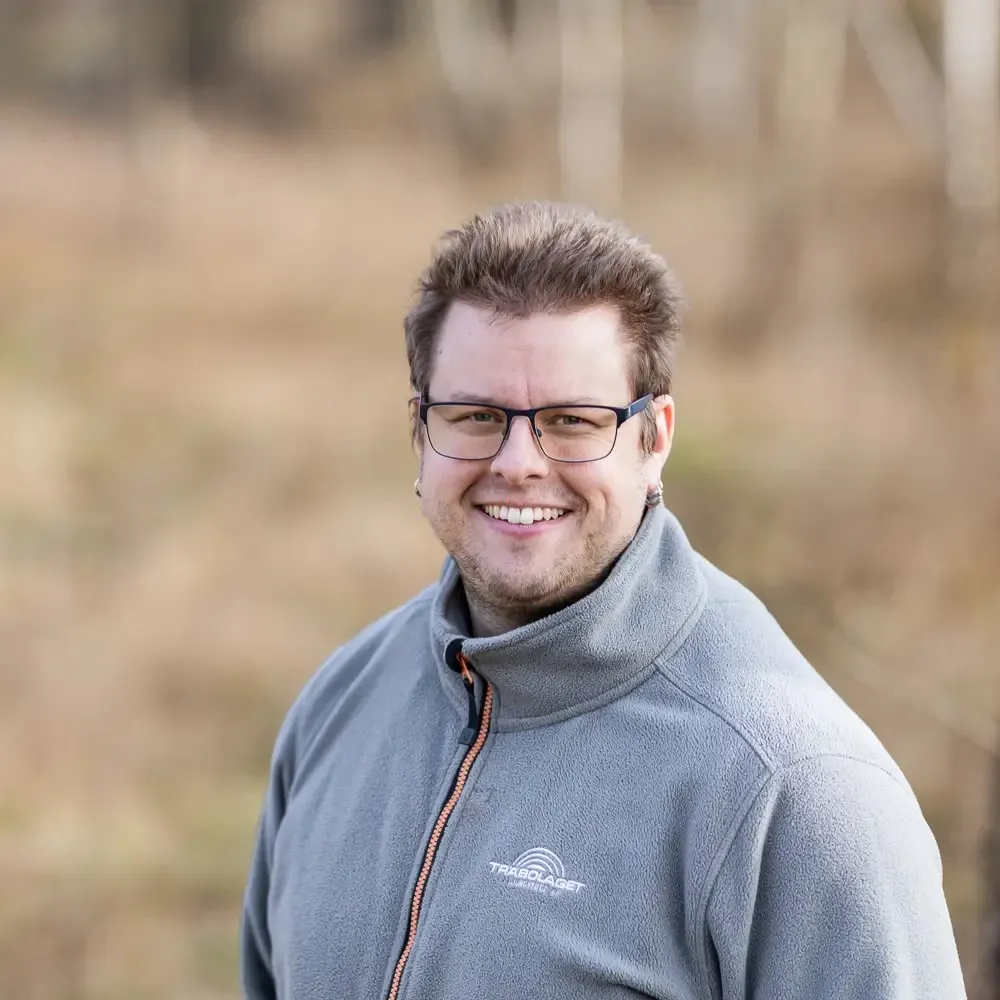 En leende man i glasögon och grå kavaj står utomhus i en suddig naturmiljö.