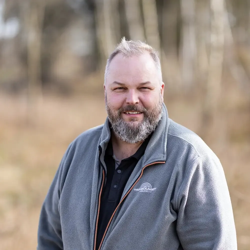 En skäggig man i grå kavaj står utomhus med träd i bakgrunden.