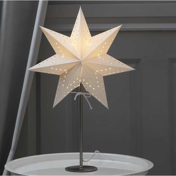Lampan Star Trading Stjärna på fot Bobo lyser vitt på ett runt bord framför en grå vägg.