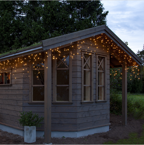 Liten trästuga med Star Trading Istappsslinga Shiny Warm White-lampor, omgiven av träd.